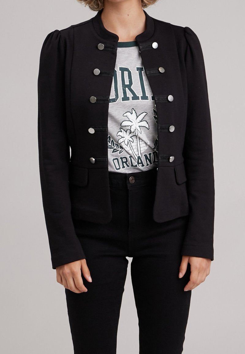 Veste noire cintrée avec épaules bouffantes, boutons-pression argentés et texture lisse, portée sur un t-shirt graphique gris et un jean noir.