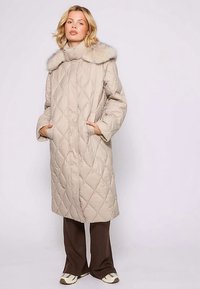 Beżowy pikowany długi płaszcz z wysokim kołnierzem i ozdobą z futerka syntetycznego. Posiada boczne kieszenie oraz smukłą sylwetkę. Miękka, podszyta tekstura.
