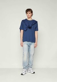 Jauns vīrietis stāv, uzvilcis tumši zilu T-kreklu ar atbilstošu džemperi, kas atdarināts uz pleciem, gaiši zilas džinsus un baltas sporta apavus.