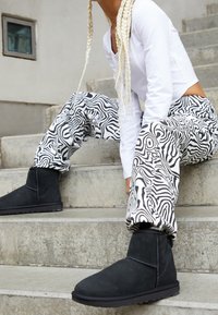 Czarne zamszowe botki za kostkę o teksturowanym wykończeniu, w zestawieniu z białą bluzką z długim rękawem oraz spodniami z czarno-białym abstrakcyjnym wzorem.