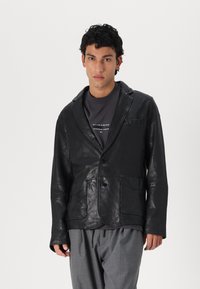 Schwarze Lederjacke mit Reverskragen, zwei Vorderstaschen und Knopfverschluss. Getragen über einem dunkelgrauen Hemd und lockeren grauen Hosen.