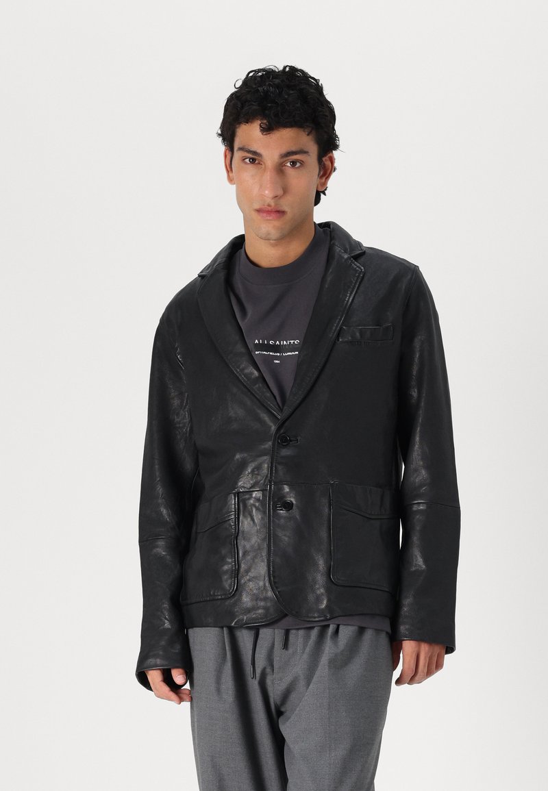 Schwarze Lederjacke mit Reverskragen, zwei Vorderstaschen und Knopfverschluss. Getragen über einem dunkelgrauen Hemd und lockeren grauen Hosen.