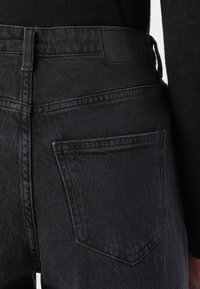 Κοντινή λήψη από την πίσω τσέπη και τη ζώνη μαύρων τζιν με ραμμένες λεπτομέρειες και δερμάτινη μαύρη ετικέτα.