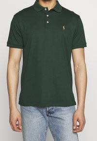 Zelená bavlněná polo košile s klasickým límcem, krátkými rukávy a třemi knoflíky. Obsahuje malé vyšívané logo na hrudi.