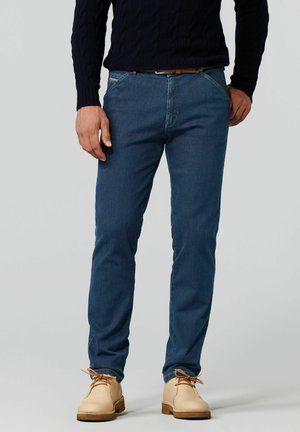 Jean bleu foncé avec une coupe slim, comportant des poches avant et des coutures discrètes. Porté avec des chaussures beiges et un pull en maille torsadée bleu marine.
