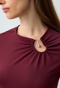 Burgundowa bluzka z okrągłym dekoltem, z marszczonym materiałem i złotą ozdobą przy szyi, prezentująca gładką teksturę.