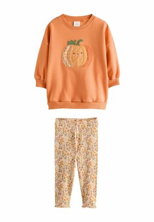 Orangefarbener Langarm-Kinderpullover mit einem lachenden Kürbis-Design, kombiniert mit beigen Leggings mit Blumenmuster.