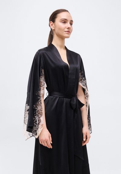 Γυναίκα που φοράει μαύρο σατέν ρόμπα με διάφανα φαρδιά μανίκια από λουλουδάτη δαντέλα, δεμένη στη μέση με μια αντίστοιχη σατέν ζώνη.