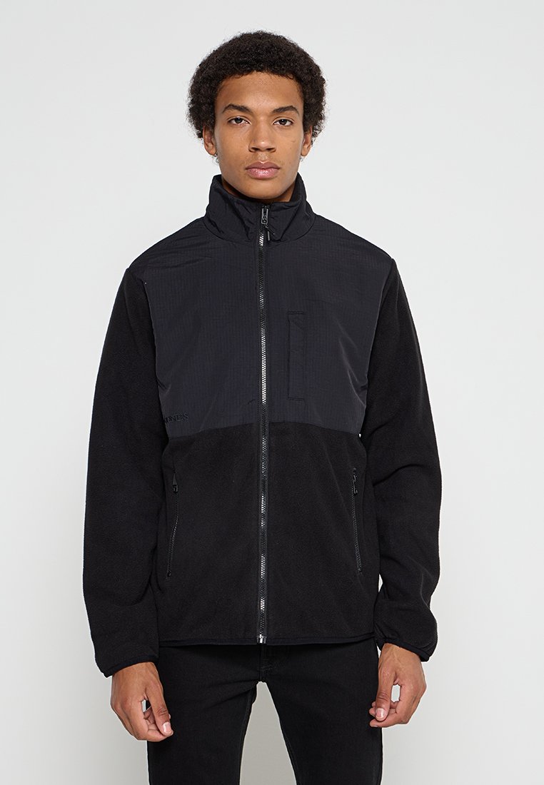 Veste en polaire noire avec un panneau supérieur texturé, un col montant et des poches zippées. Elle présente un devant lisse et des poignets élastiques.