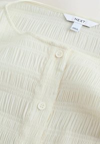 Skjorte i lys creme med horisontal rynkekonstruktion, der har to knapper og en afrundet halsudskæring. Mærkeetiket synlig.