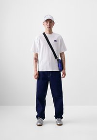 T-shirt branca com um pequeno emblema, calças de ganga azul marinho de corte largo, ténis prateados e um boné prateado. Apresenta uma mochila transversal azul.