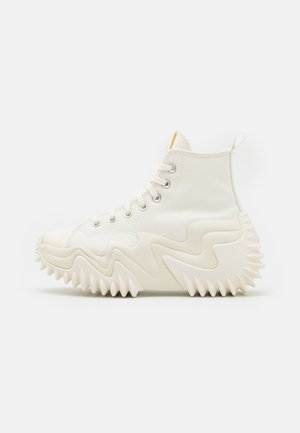 Hoge off-white sneaker met dikke, gekartelde zigzagzool en stoffen bovenwerk met metalen oogjes voor veters, gezien vanaf de zijkant.