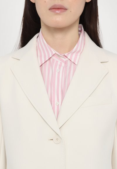 Blazer beige chiaro con un solo bottone e rever a punta, indossato sopra una camicia colletto a righe rosa e bianche. Texture liscia, dettagli minimali.