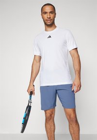 Bela kratka športna majica s subtilno teksturo in črnim logotipom, v kombinaciji z modrimi kratkimi hlačami. Drži črno-modro teniško lopar.