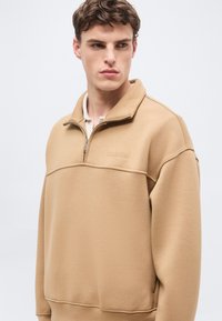 Νεαρός με κοντά καστανά μαλλιά που φοράει μπεζ φούτερ με φερμουάρ μέχρι το στήθος και διακριτικό κεντημένο λογότυπο, κοιτάει προς τα πλάγια με φόντο ένα απλό φόντο.