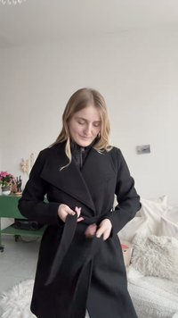 Juodas, ilgomis rankovėmis paltas su įvyniojamu dizainu. Turi aukštą apykaklę ir surišamą diržą. Lygus tekstūros paviršius, minimalistinis stilius, be matomų detalių.