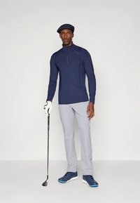 Marineblå golf-pullover med lange ermer og glidelås, grå bukser og marineblå sko. Modellen holder en golfkølle og har på seg en flat cap og golfhanske.