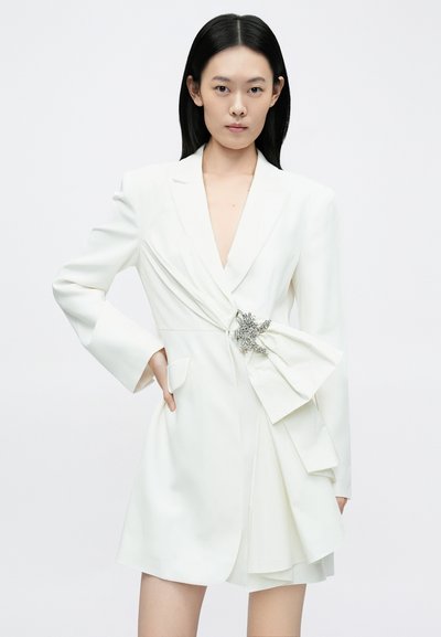 Vestido blazer branco com decote em V profundo e corte ajustado. Apresenta um detalhe estruturado de laço na cintura adornado com aplicações prateadas.