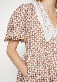 Femme portant une robe blanche avec des motifs géométriques roses et orange, un col en dentelle, des manches bouffantes, et les mains dans les poches latérales.
