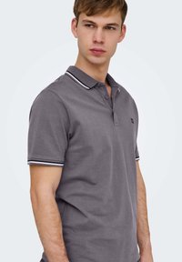 Ung mand iført grå kortærmet polo med sort-hvid stribet krave og ærmekanter, kigger til siden.