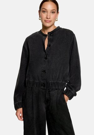 Femme portant une veste noire boutonnée à manches longues avec taille élastique et pantalon noir assorti, debout avec une main dans la poche.