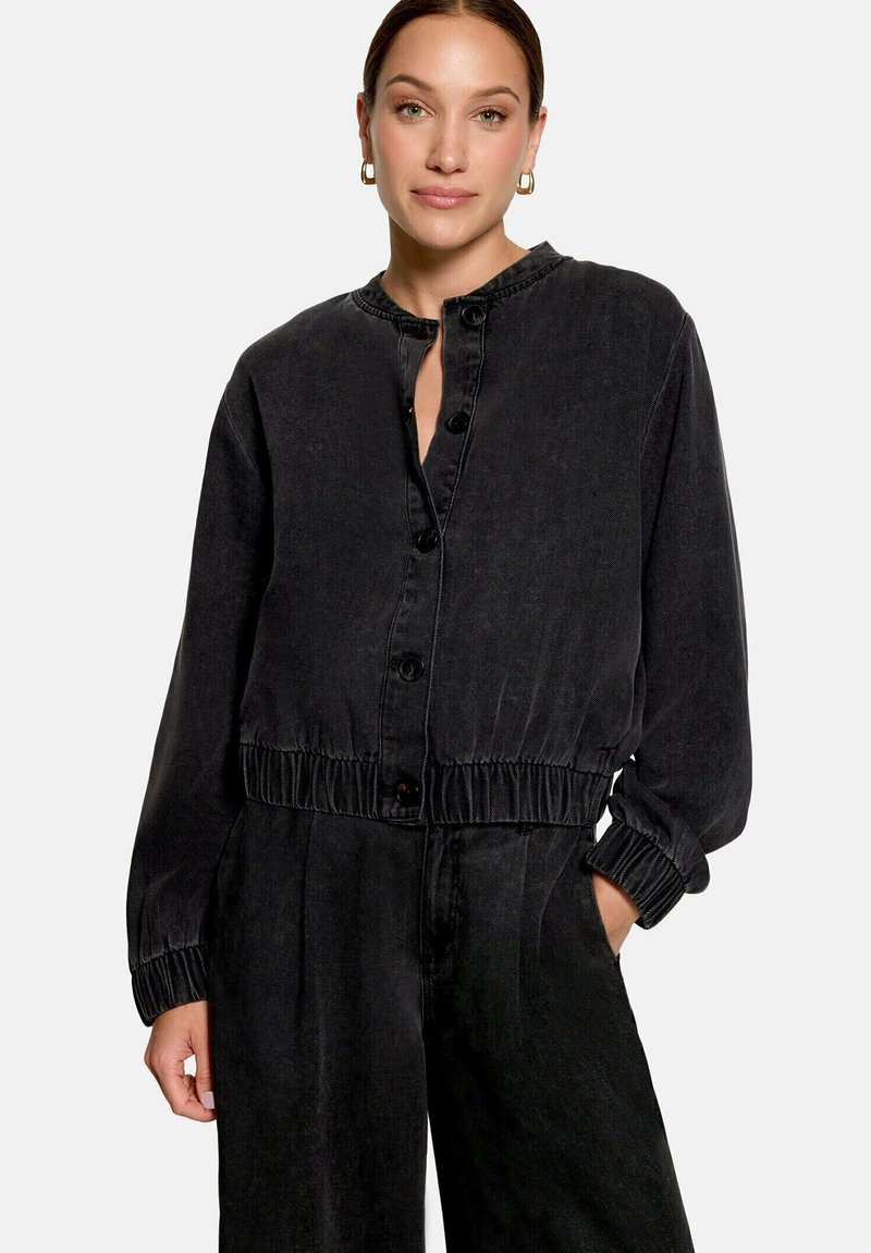 Femme portant une veste noire boutonnée à manches longues avec taille élastique et pantalon noir assorti, debout avec une main dans la poche.