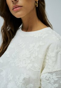 Suéter blanco texturizado con bordado floral, lentejuelas y un escote redondo, combinado con pendientes de aro dorados.