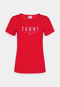 Czerwona bawełniana koszulka z dekoltem w serek, z wyraźnym białym logo "TOMMY JEANS" oraz małą grafiką flagi z przodu. Krótkie rękawy, luźny krój.
