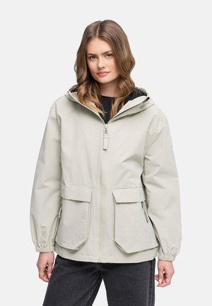 Tânără purtând o geacă deschisă la culoare, gri, cu glugă, buzunare mari frontale și manșete elastice, stând pe un fundal simplu.