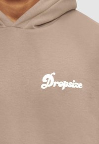 Hettegenser i lys beige bomull med en myk tekstur. Har en hvit brodert logo "Dropsize" på forsiden.