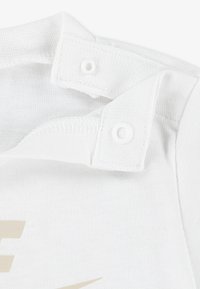 Balto medvilninė marškinėlių apykaklė su spaudžiamais užsegimais ir banguota tekstūra; po ja yra auksinės spalvos logotipo grafika.