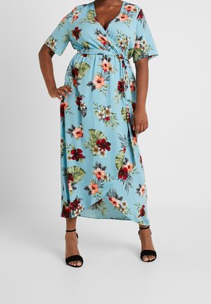 Žena oblečená v lehkých modrých kvetovaných zavinovacích šatech s krátkými rukávy a černými kotníkovými botami na podpatku, stojící s jednou rukou na boku.