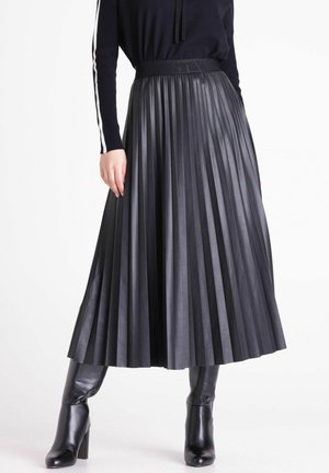 Vrouw die een zwarte geplooide midi-rok draagt, een zwart top met lange mouwen met een witte streep op de mouw en zwarte laarzen met hak tegen een witte achtergrond.