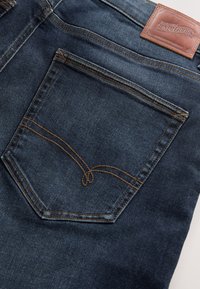 Tasca posteriore di jeans denim blu scuro con cuciture marroni e patch in pelle color beige con etichetta "Premium Denim Goods."