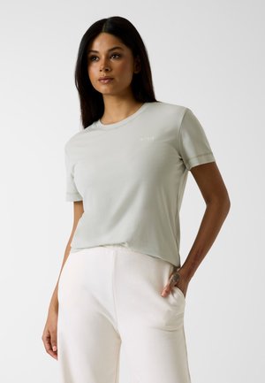 Frau mit langen dunklen Haaren, die ein hellgraues T-Shirt mit kurzen Ärmeln und eine weiße Hose trägt, steht mit einer Hand in der Tasche vor einfarbigem Hintergrund.