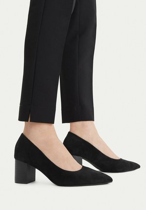 Osoba nosící černé semišové lodičky s nízkým blokovým podpatkem, spojené s černými zkrácenými kalhotami s rozparky vpředu na lemech.