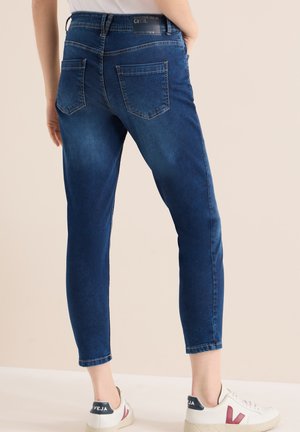 Femme portant un jean skinny bleu foncé coupé court avec des poches arrière et des baskets blanches Veja, debout devant un fond beige uni.
