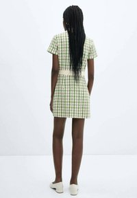 Vestito corto con motivo a quadretti verdi e bianchi, maniche corte, vita aderente e tessuto strutturato. Indossato con scarpe di colore chiaro.