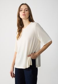 Lys kremfarget bluse laget av myk stoff, med rund halslinning, ermelange ermer og sidelommer, parret med mørke bukser.