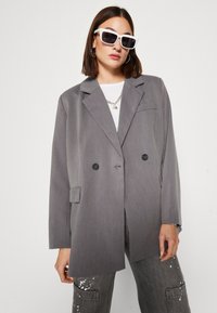 Grå dubbelknäppt blazer i texturerat tyg, med slagkrage, två svarta knappar och sidofickor.