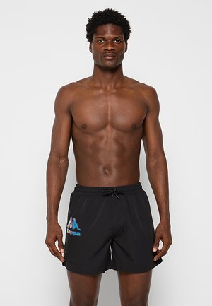 Uomo senza maglietta con pelle scura e capelli corti e ricci che indossa pantaloncini da bagno neri Kappa con logo sulla coscia sinistra, in piedi davanti a uno sfondo semplice.