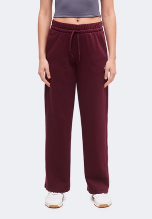Persoon die bordeaux sweatpants met trekkoord draagt, een grijs cropped topje en witte sneakers, staand tegen een effen achtergrond.