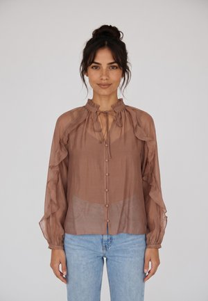 Donna con i capelli scuri raccolti che indossa una camicetta marrone trasparente a maniche lunghe con volant e jeans azzurri, in piedi davanti a uno sfondo semplice.