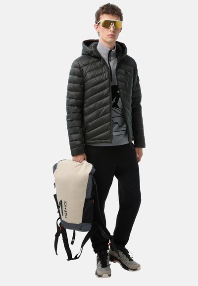 Daunenjacke in Dunkelgrün, grauer Rollkragenpullover, schwarze Hose und graue Sneakers. Hält einen cremefarbenen und grauen Rucksack mit Logo.