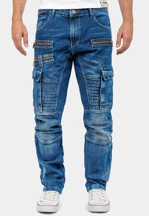 Pantalons cargo en denim bleu avec de multiples poches zippées et des panneaux texturés. Possède une jambe fuselée et des coutures visibles. Portés avec des baskets blanches.
