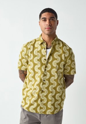 Moški z kratkimi lasmi in tetovažami, oblečen v rumeno-oker majico s kratkimi rokavi in belimi okroglimi vzorci ter svetlo sive hlače.