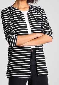 Black and white striped cardigan with three-quarter sleeves, open front, and a soft, textured knit material. Worn over a white top.