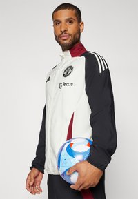 Witte en zwarte sportjacket met bordeauxrode accenten, featuring een logo en de tekst "Tezos". Model houdt een meerkleurige voetbal vast.