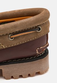 Chaussure en daim et cuir marron avec un col rembourré, des coutures marron, des œillets en métal et une semelle en caoutchouc texturée.