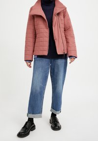 Prošívaná, světle růžová bunda s prošívaným vzorem a širokým límcem, nošená přes tmavě modrý rolák; doplněná volnými modrými džínami a černými botami.